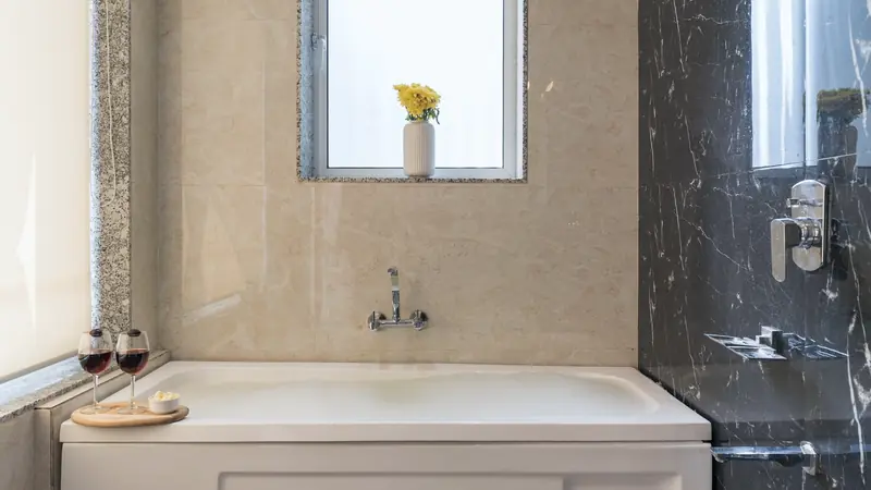 Baignoire blanche à tablier devant un mur en grands carreaux beiges effet marbre, sous une petite fenêtre