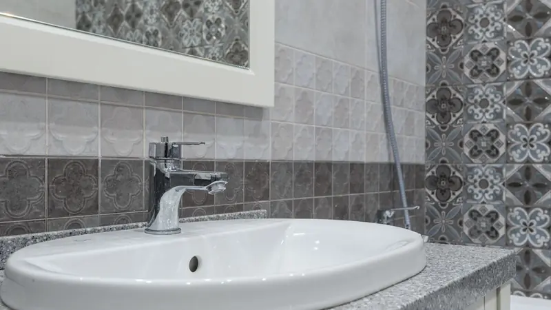Vasque blanche encastrée dans un plan gris moucheté, devant un carrelage gris en relief et une bande de carreaux à motifs floraux bleus et gris