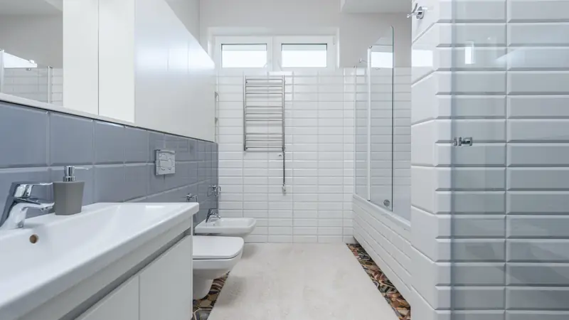 Salle de bain étroite carrelée de blanc en carreaux biseautés, tablier de baignoire carrelé et paroi de douche vitrée
