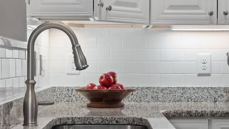 Crédence en carreaux blancs rectangulaires au-dessus d'un plan de travail en granit, avec évier inox et prises murales