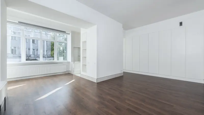 Appartement vide et lumineux avec parquet foncé, grande fenêtre, radiateur blanc et placards intégrés