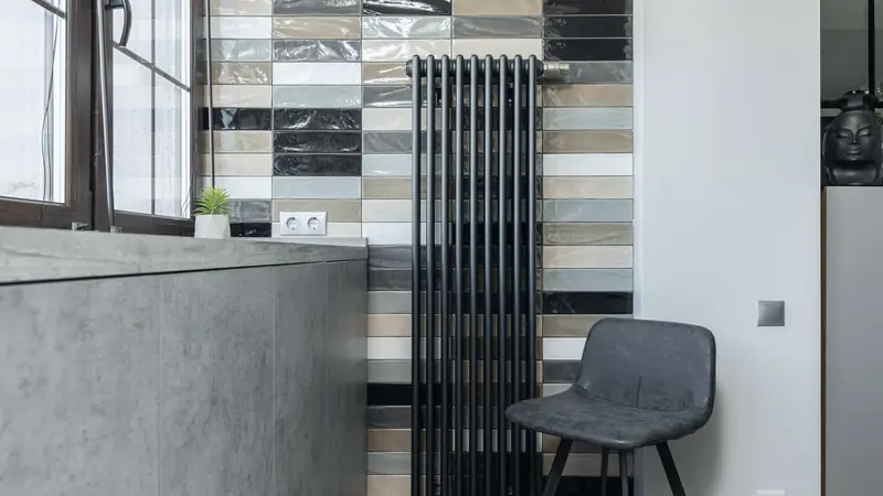 Radiateur vertical noir à colonnes contre un mur de carreaux vernissés, près d'une fenêtre