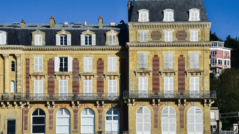 Façade d'un grand immeuble ancien en briques jaunes et rouges, aux volets blancs, balcons en fer forgé et toit d'ardoise