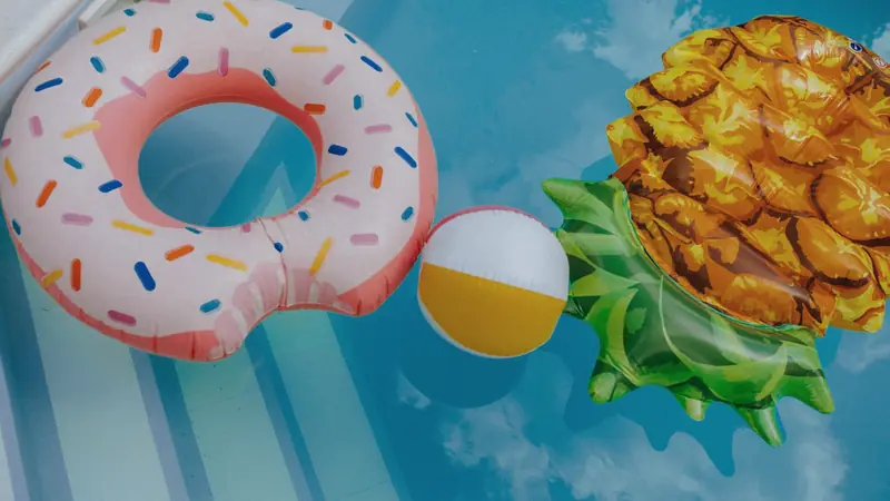 Bouée beignet rose, ballon de plage blanc et jaune et matelas en forme d'ananas flottant près du bord d'une piscine