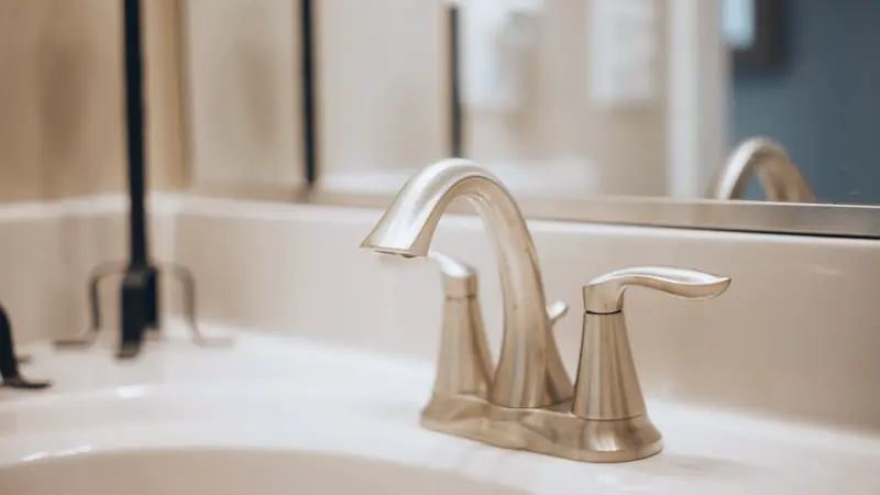 Robinet mélangeur à deux manettes en finition nickel brossé sur une vasque blanche, miroir en arrière-plan