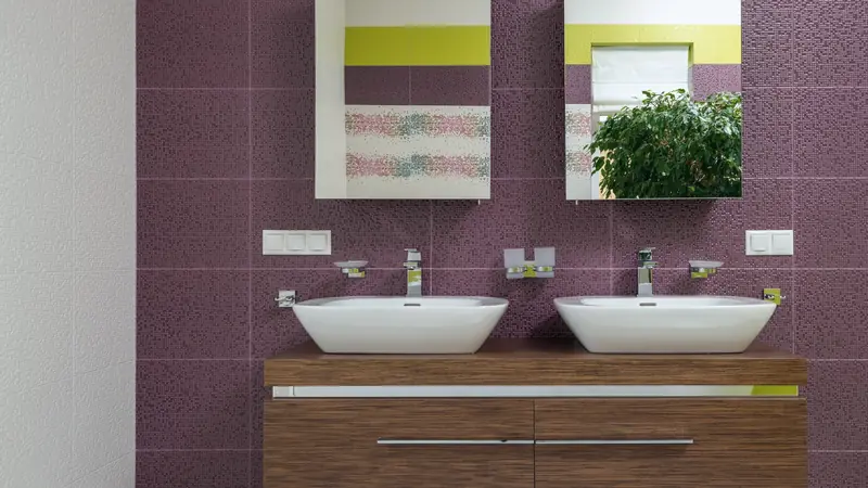 Meuble suspendu en bois à deux tiroirs portant deux vasques blanches à poser, devant un mur de mosaïque violette et deux miroirs