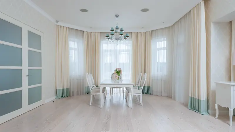 Salle à manger lumineuse au parquet de bois clair à larges lames, avec une table blanche devant des fenêtres voilées
