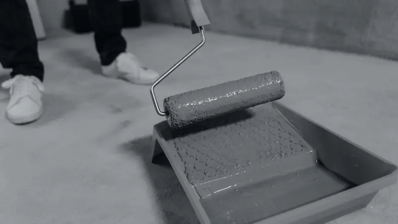 Rouleau de peinture chargé au-dessus d'un bac posé sur un sol en béton, photo en noir et blanc