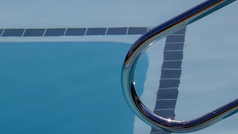 Main courante en inox d'une échelle de piscine au-dessus d'une eau bleue et d'une ligne de carreaux sombres