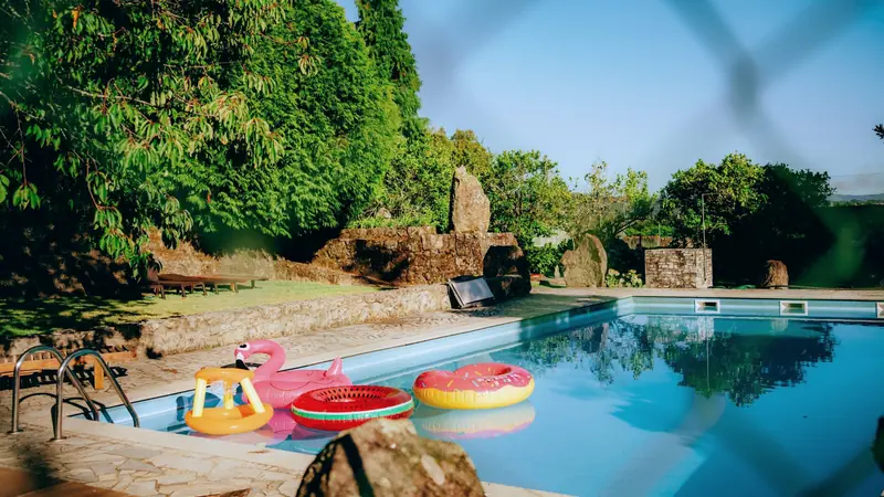 Piscine de jardin bordée de dalles de pierre, avec des bouées colorées, vue à travers un grillage flou au premier plan