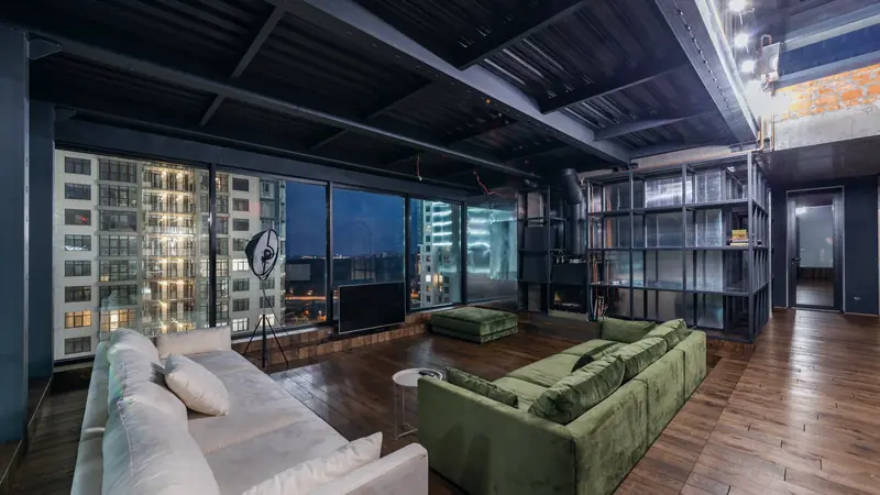 Grand séjour de loft aux baies vitrées à cadres noirs donnant sur des immeubles éclairés le soir
