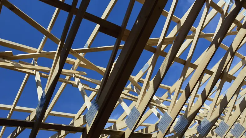 Fermettes de charpente en bois clair assemblées par des connecteurs métalliques, vues d'en dessous sur fond de ciel bleu
