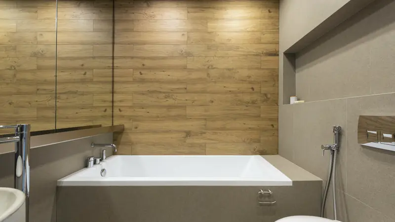 Baignoire droite blanche encastrée avec tablier carrelé taupe, mur en carreaux imitation bois et robinetterie sur le rebord
