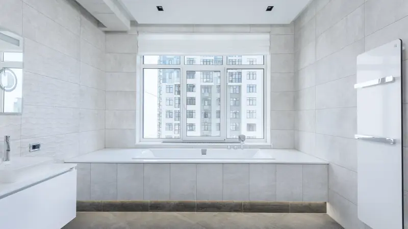 Baignoire rectangulaire encastrée dans un coffrage carrelé gris clair, sous une large fenêtre, avec sèche-serviettes blanc