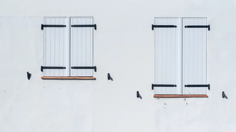 Façade enduite blanche avec deux fenêtres aux volets blancs fermés et ferrures noires